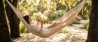 Young woman relaxing in hammock under trees, reading book in sunny park. Leisure, vacation, outdoor lifestyle concept for wellness and relaxation designs.