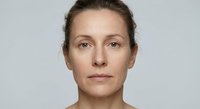 Natural portrait of middle-aged Caucasian woman with brown hair pulled back, neutral expression, clean skin, professional headshot on gray background.