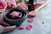 Spa wellness setup with pink roses, sea salt scrub in bowl, essential oil bottle and wooden spoon for natural skincare and aromatherapy relaxation.