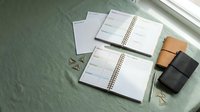 Organized workspace with open planners and notebooks on green textured surface near window for productivity and scheduling tasks.