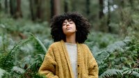 Young woman with curly hair wearing mustard cardigan standing in lush forest with ferns, eyes closed in peaceful meditation moment.