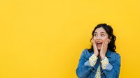 Young woman with dark wavy hair and medium skin tone wearing denim shirt, expressing surprise and excitement with hands on face against bright yellow background.