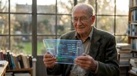 Senior Caucasian man with glasses examining transparent digital interface screen in modern office environment with natural lighting and bookshelves.