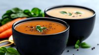 Fresh homemade creamy soup in black ceramic bowls garnished with herbs and spices, served with raw carrots and fresh basil leaves on dark background.