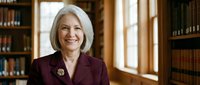 Professional mature Caucasian businesswoman with gray hair smiling confidently in burgundy blazer standing in elegant library office with wooden bookshelves.