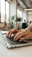 Young woman typing on laptop keyboard in bright modern office, colleagues in background, close-up of hands, productive workspace.