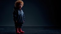 Young child with curly hair wearing leather jacket and red rain boots standing on dark background, ready for rainy weather adventure.