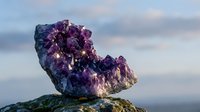 Sparkling amethyst crystal cluster on a rock against a soft blue sky, showcasing deep purple gemstone formations with glowing light reflections.