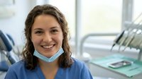 Young female dentist with long brown wavy hair wearing blue scrubs and surgical mask smiling at camera in modern dental clinic office setting with equipment visible.