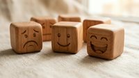 Wooden blocks with carved emoji faces showing happy, sad, and laughing expressions arranged on linen fabric, representing emotions and feelings.