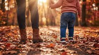 Parent and toddler walking together on autumn leaves path during golden hour sunset creating warm family bonding moment in fall season park setting.