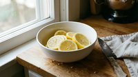 Fresh lemon slices in white ceramic bowl on rustic wooden cutting board by kitchen window with natural lighting for healthy cooking and food preparation.