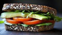 Whole grain seeded bread sandwich with fresh cucumber, tomato, and leafy greens on dark surface for food and nutrition content.