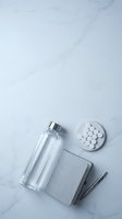 Clear glass bottle with silver cap, white pills, gray notebook and pen arranged on white marble surface for healthcare and wellness concepts.