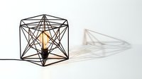 Geometric wire frame table lamp with Edison bulb casting intricate shadow patterns on white background, modern industrial interior decor.