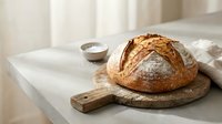 Freshly baked artisan sourdough bread loaf on wooden cutting board in bright kitchen setting with natural lighting for food photography and cooking content.