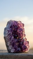 Sparkling amethyst crystal cluster glowing in sunlight, purple gemstone geode on wooden surface with soft sky background.