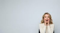 Excited young woman with blonde wavy hair wearing cream blazer, expressing surprise and joy with open mouth and hands near face against neutral gray background.