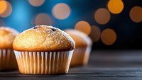 Fresh baked vanilla muffin with sugar powder on dark wooden table against blurred bokeh lights background, selective focus on pastry dessert.