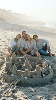 Happy family of four posing together at sandy beach with sandcastle during golden hour sunset, enjoying outdoor leisure time and vacation bonding moments.
