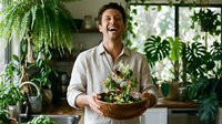 Cheerful young Caucasian man laughing while tossing fresh salad in wooden bowl, surrounded by lush indoor plants in bright kitchen.