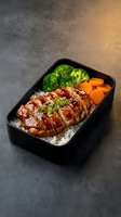 Glazed teriyaki chicken bento box with steamed broccoli and carrots over white rice in black container for healthy meal prep and Japanese cuisine.