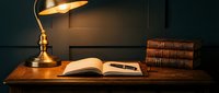 Vintage brass desk lamp illuminating open book and fountain pen on wooden desk with leather-bound books in warm atmospheric study setting for reading.