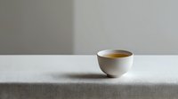 White ceramic cup filled with golden tea on minimalist white textured tablecloth against neutral gray wall background, creating serene morning atmosphere.