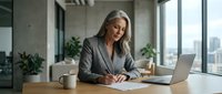 Confident mature Caucasian businesswoman with gray hair signing documents at office desk with laptop, embodying leadership and professionalism.
