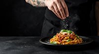Chef with tattoos seasoning spaghetti bolognese pasta dish with fresh basil garnish on dark plate against black background for culinary content.