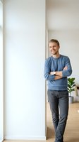 Young Caucasian male professional standing confidently with crossed arms in modern bright office space, wearing casual blue sweater and dark pants for corporate portraits.