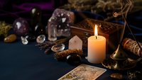 Mystical fortune telling setup with burning candle, tarot cards, crystals, amethyst geode and brass scales on dark surface for divination practices.