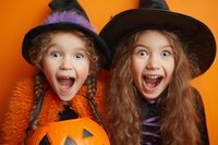 Two excited young girls in Halloween costumes with witch hats and pumpkin basket showing joyful expressions against orange background, ready for trick or treating.