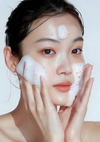 Young Asian woman applying foaming cleanser on face during skincare routine, close up portrait on white background showing fresh glowing skin.