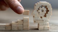 Hand placing wooden block on steps leading to gear-shaped head figure, symbolizing business growth and cognitive development process.