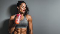 Athletic young woman drinking protein shake after workout in gym wearing gray sports bra with dramatic lighting for fitness and nutrition concepts.