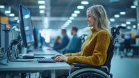 Young blonde woman in yellow sweater using computer while sitting in wheelchair at modern office workspace, showing workplace diversity and inclusion.