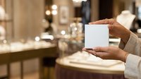 Hands holding blank white card in luxury retail store environment with blurred background, suitable for product mockups and promotional campaigns.