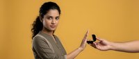 Young Hispanic woman in olive green shirt making stop gesture refusing smartphone offered by hand against yellow background for digital detox concept.