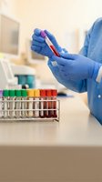 Medical laboratory technician in blue gloves examining blood sample test tube with colorful specimen collection rack in modern clinical research facility.