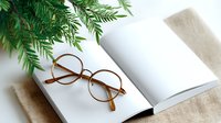 Round vintage eyeglasses on open blank book with green fern leaves on white background, minimalist composition for reading and relaxation concept.