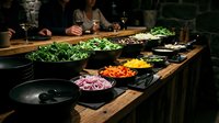 Chefs prepare fresh vegetables at wooden counter with colorful ingredients including greens, carrots, peppers and onions for cooking preparation.