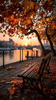 Autumn sunset over river with empty wooden bench under vibrant orange maple leaves, city skyline silhouette in background creating peaceful urban scene.