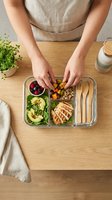 Person with light skin tone preparing a balanced meal in a compartmentalized container, featuring grilled chicken, fresh vegetables, berries, and grains for healthy eating and meal planning.