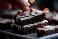 Homemade chocolate brownies decorated with white drizzle and red star sprinkles on dark plate, festive holiday baking with bokeh lights in background.