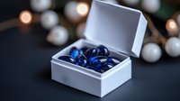 Open white gift box displaying shiny blue gemstones or decorative stones inside, surrounded by blurred pearl and gold bokeh lights on dark background for luxury product presentation.