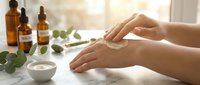 Woman applying natural skincare cream on hands with essential oil bottles and eucalyptus leaves on white table in bright sunlight for wellness routine.