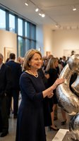 Mature Caucasian woman in navy dress examining metallic sculpture at contemporary art gallery opening with crowd of visitors in modern exhibition space.