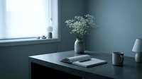 Minimalist home office desk with open notebook, ceramic vase with white flowers, mug, and lamp in soft blue-gray natural light.