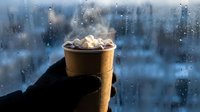Gloved hand holding steaming hot beverage cup against frosted winter window with snow-covered cityscape background. Seasonal comfort drink concept.